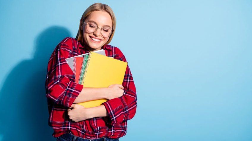 Garota loira de óculos usando blusa xadrez abraçando vários cadernos.