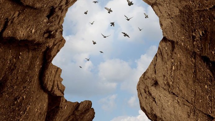 Abertura entre rochas em formato de cabeça humana, com céu e pássaros ao fundo.