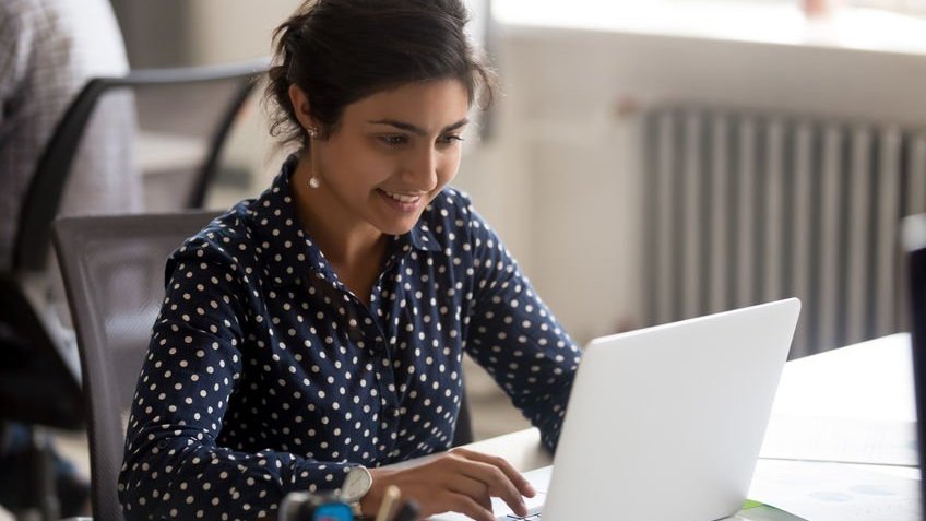 Mulher de cabelo amarrado usando camisa e sorrindo enquanto mexe no computador