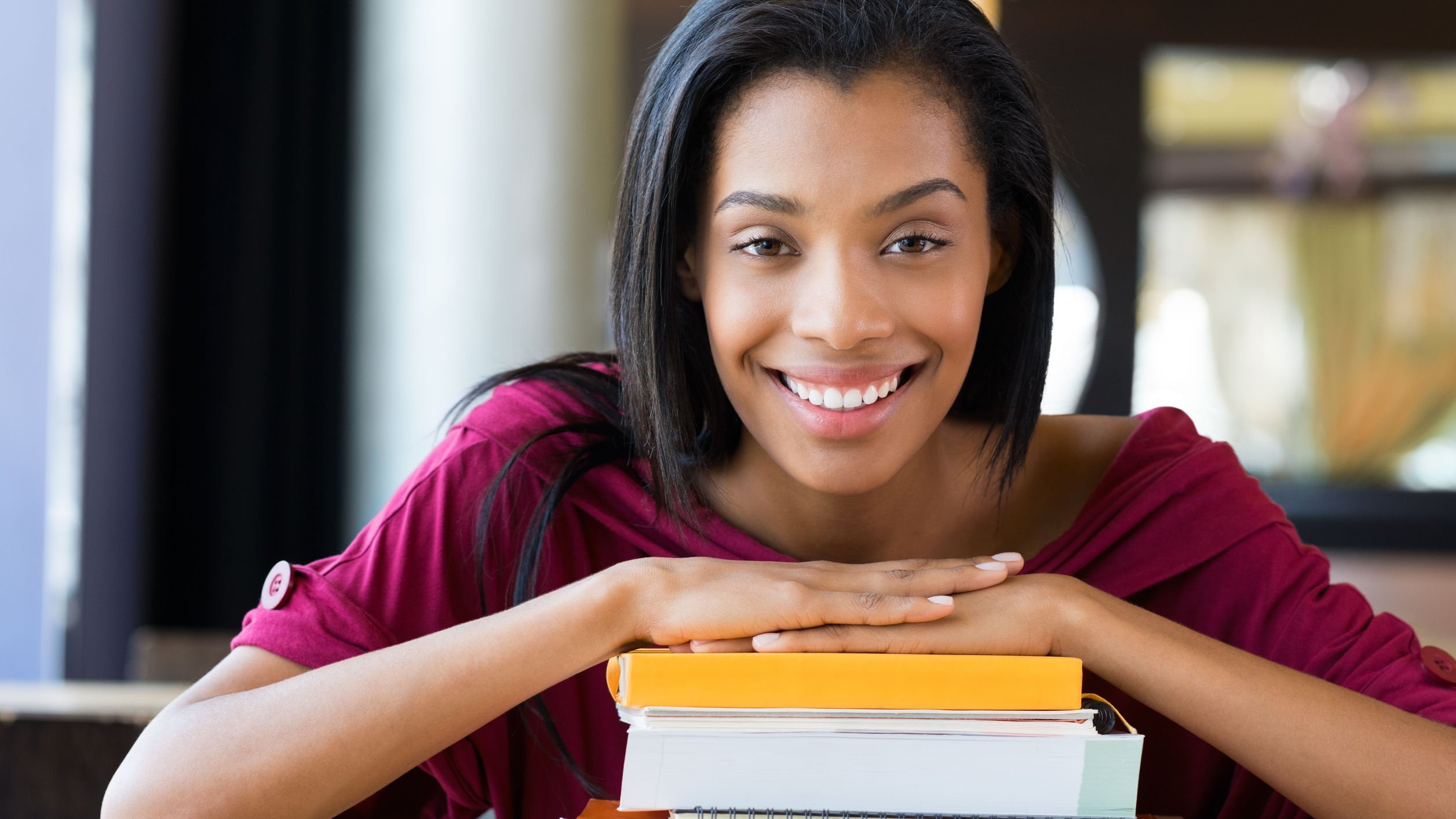 Mulher sorridente com ambas as mãos sobre uma pilha de livros e cadernos