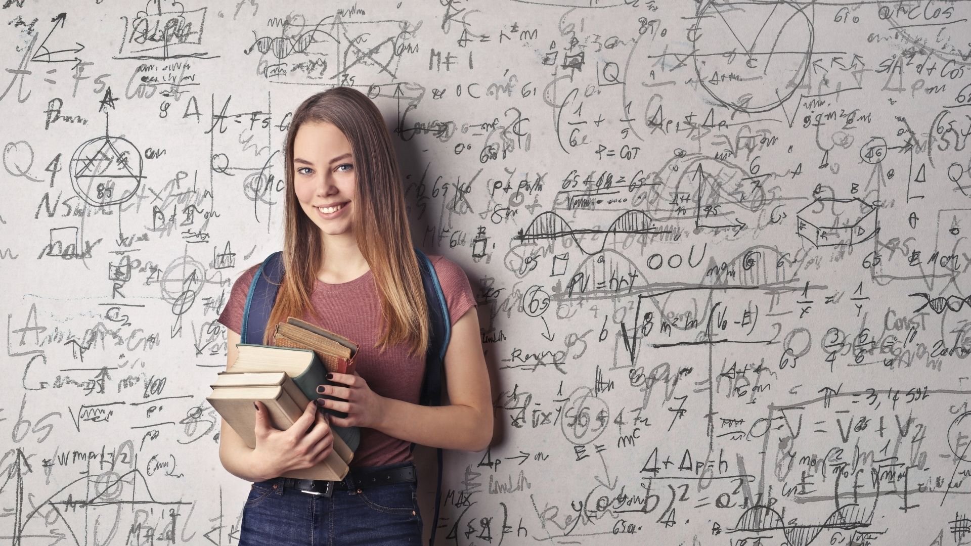Garota segurando livros em frente uma lousa preenchida com fórmulas de física