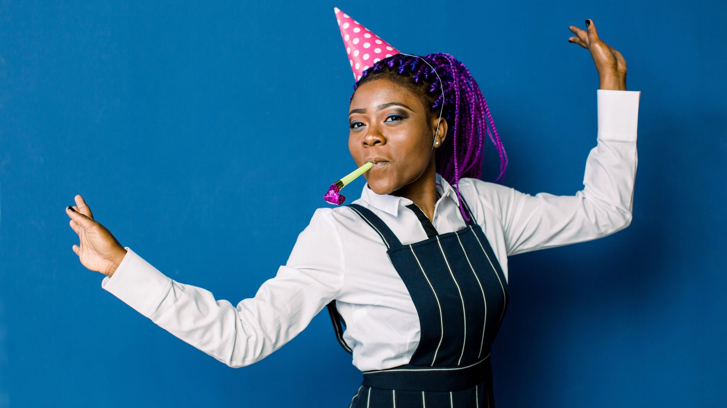 Mulher negra sorrindo, pulando com língua de sogra na boca e chapéu de festa