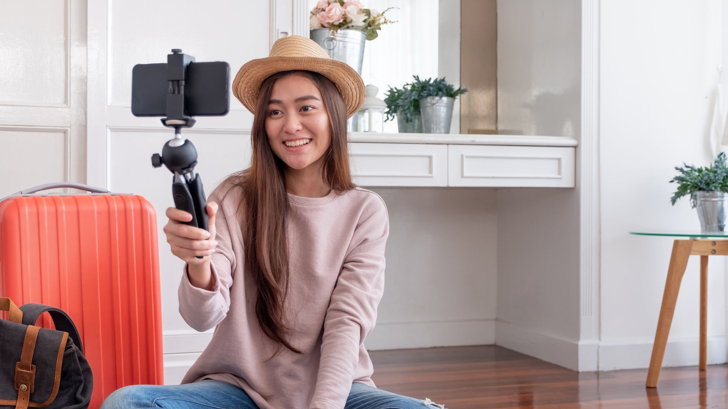 Mulher  sentada em quarto, sorrindo, segurando celular preso em pau de selfie.