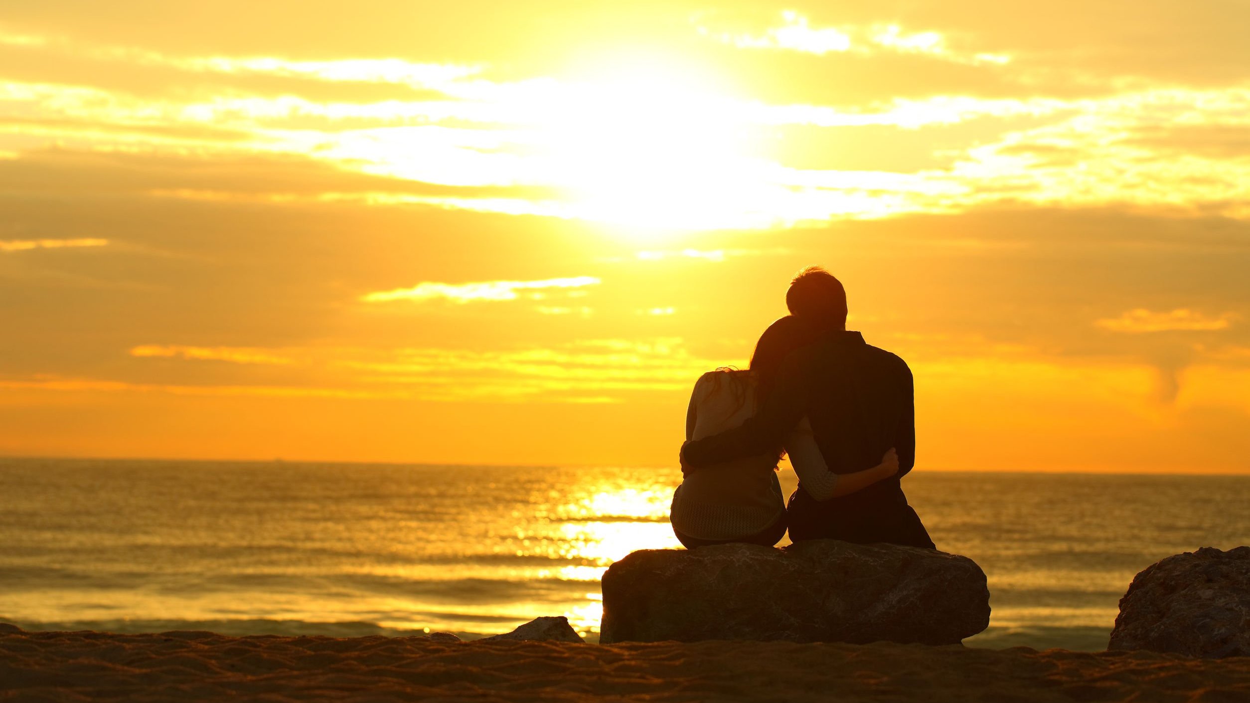 Namorados se abraçando, sentados em pedra na praia, observando por do sol.