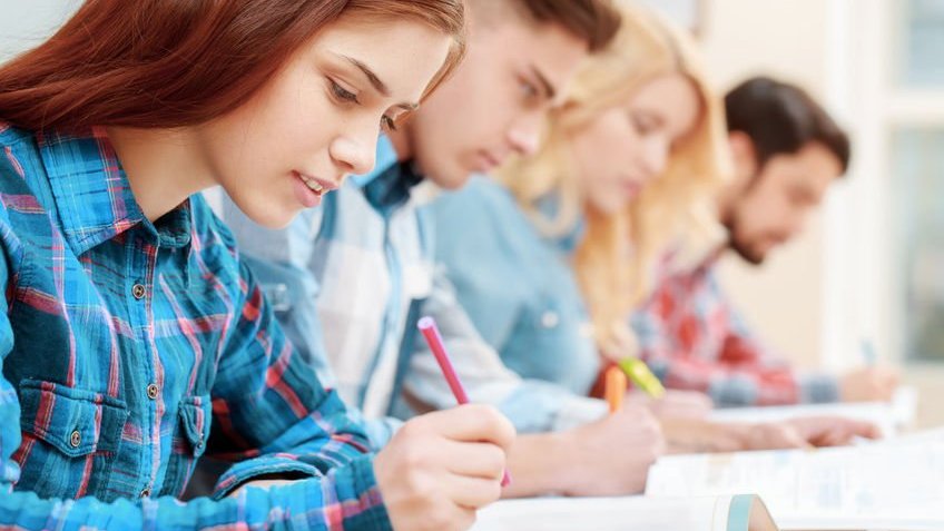 Jjovens estudantes sentados em uma fileira escrevendo.