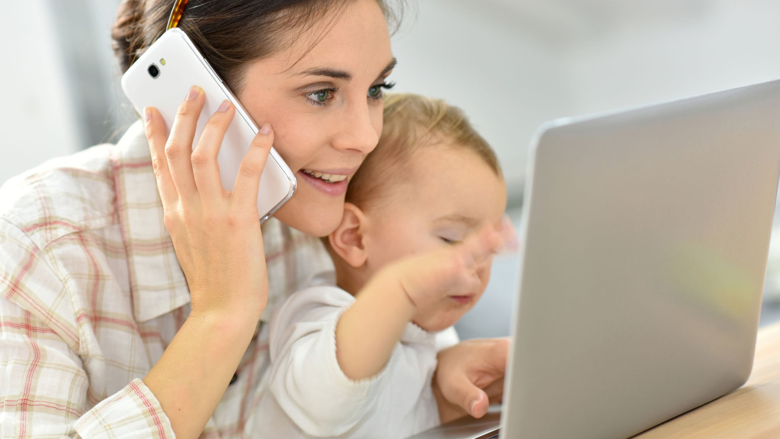 Mãe segurando filho, falando no celular em frente a notebook.