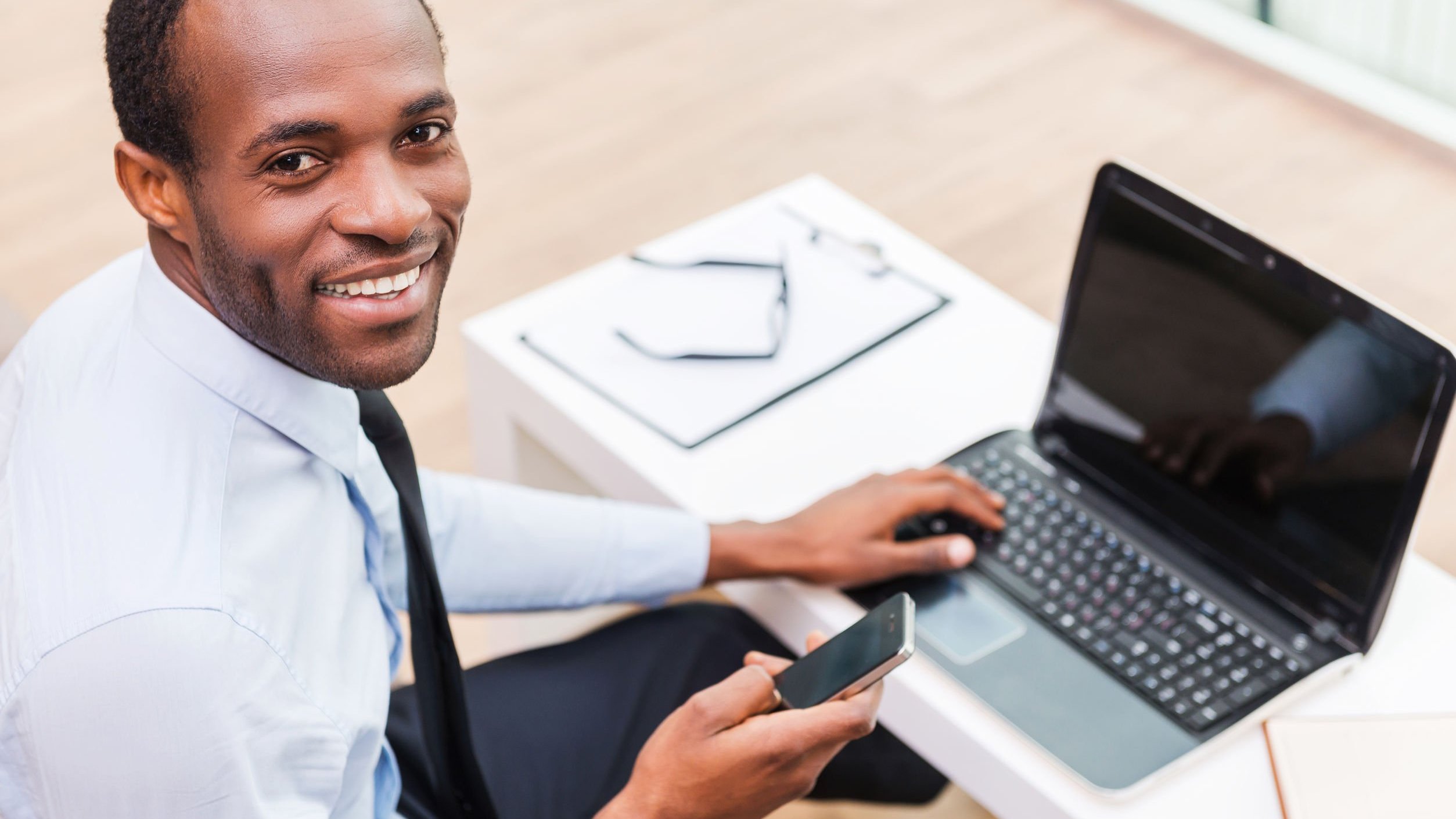 Homem negro sorrindo, segurando celular e mexendo em notebook.