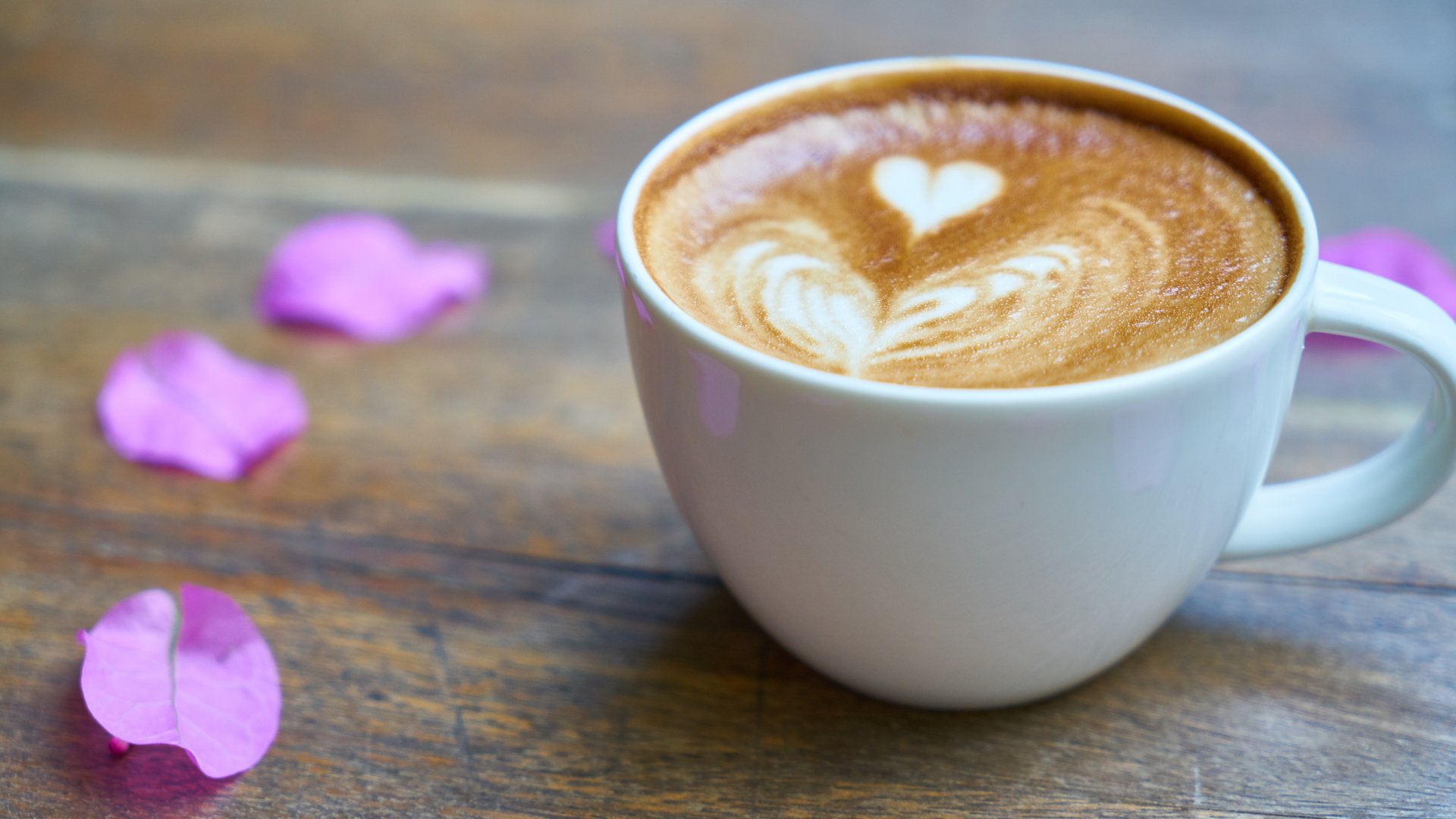 Xícara de café com desenho de coração e pétalas de flor roxa sobre mesa de madeira