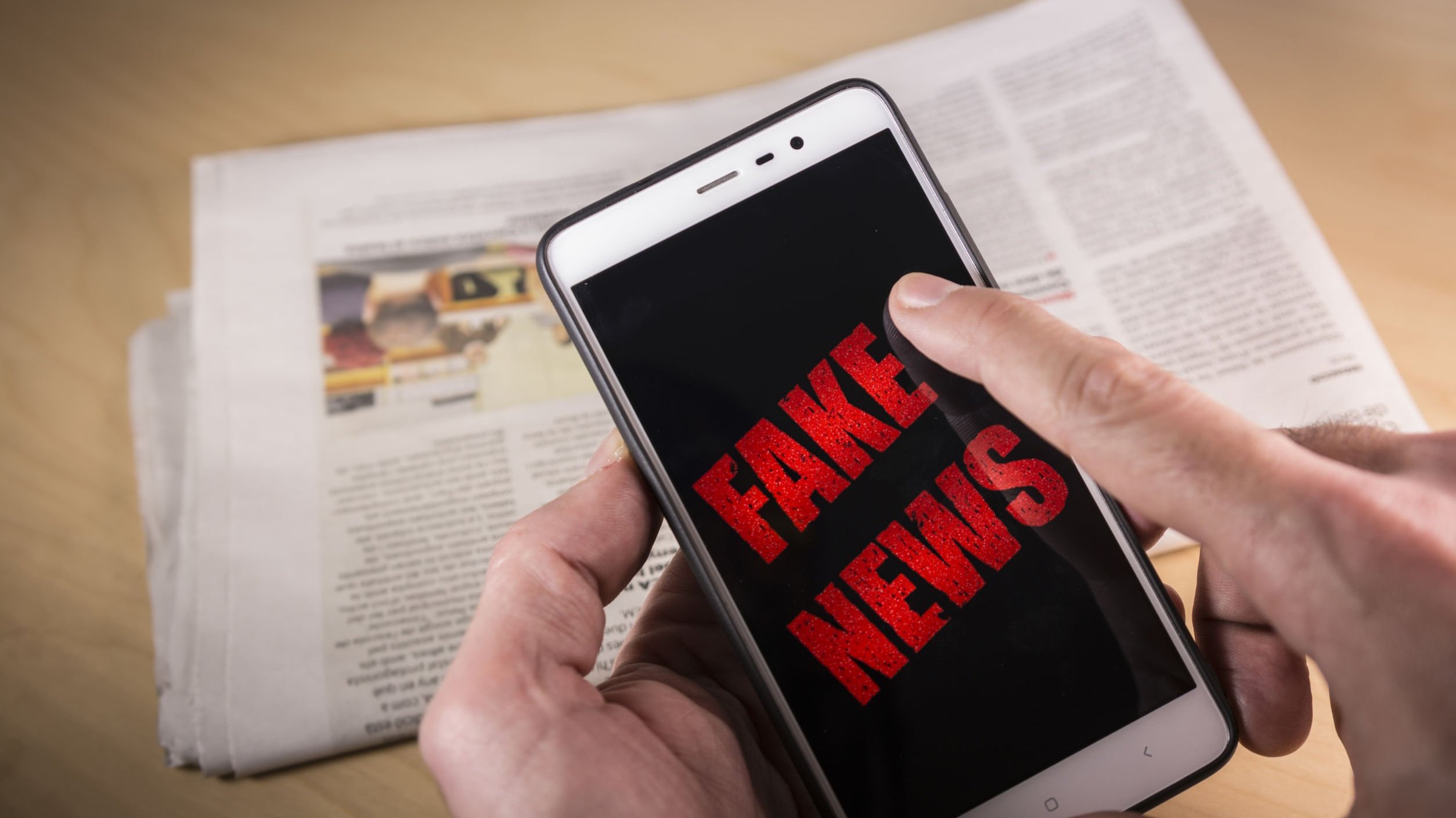 Foto de celular escrito fake news na tela, e jornal ao fundo em cima de mesa.