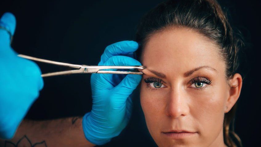 Mãos enluvadas de bodypiercer segurando ferramenta para piercing em sobrancelha