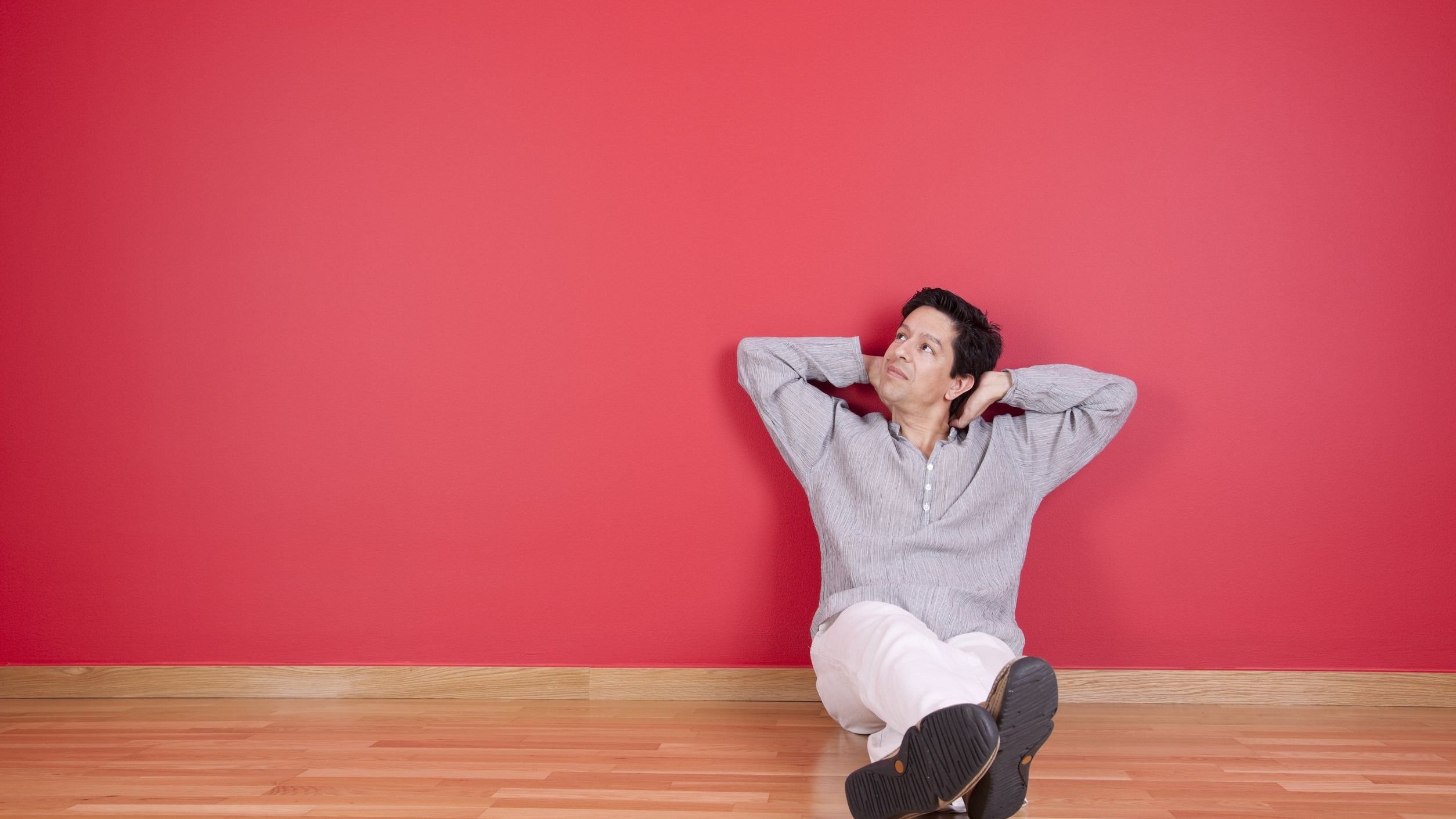 Homem sentado no chão. Ele está encostado em uma parede vermelha, olha para cima e apoia a nuca sobre as duas mãos.