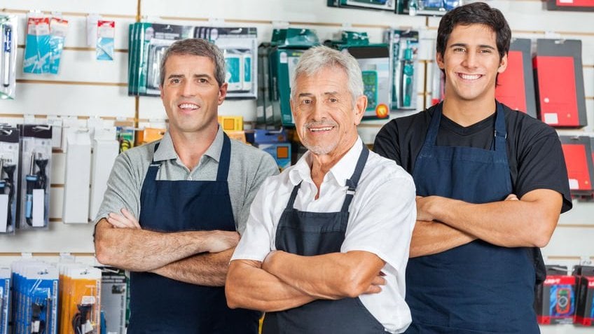 Três homens atendentes de loja posando para a foto, sorrindo e de braços cruzados