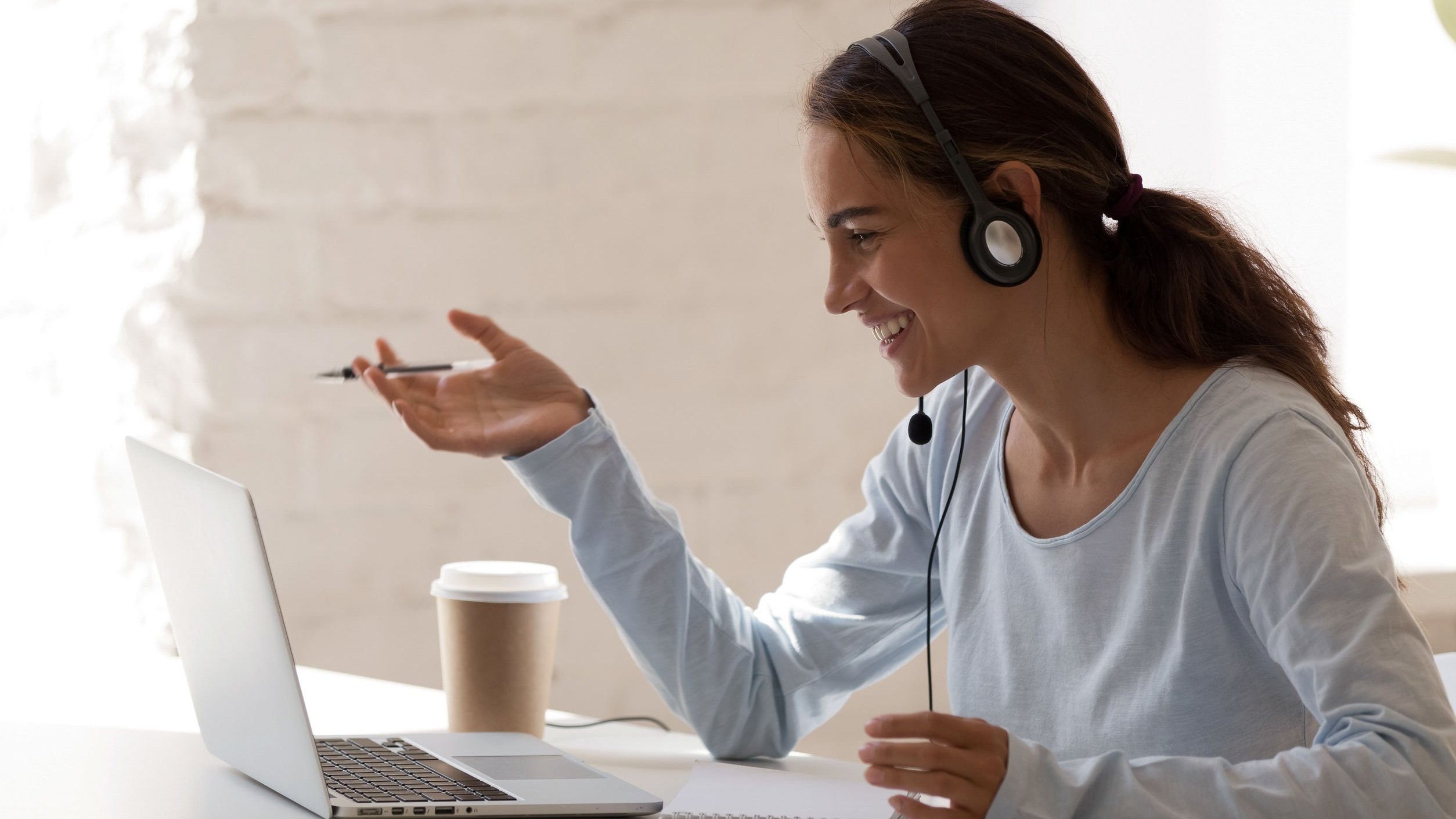 Mulher sorrindo segura caneta e usa fones de ouvido. Ela está sentada à mesa e olhando para um computador; ao lado dele, há um copo de café.