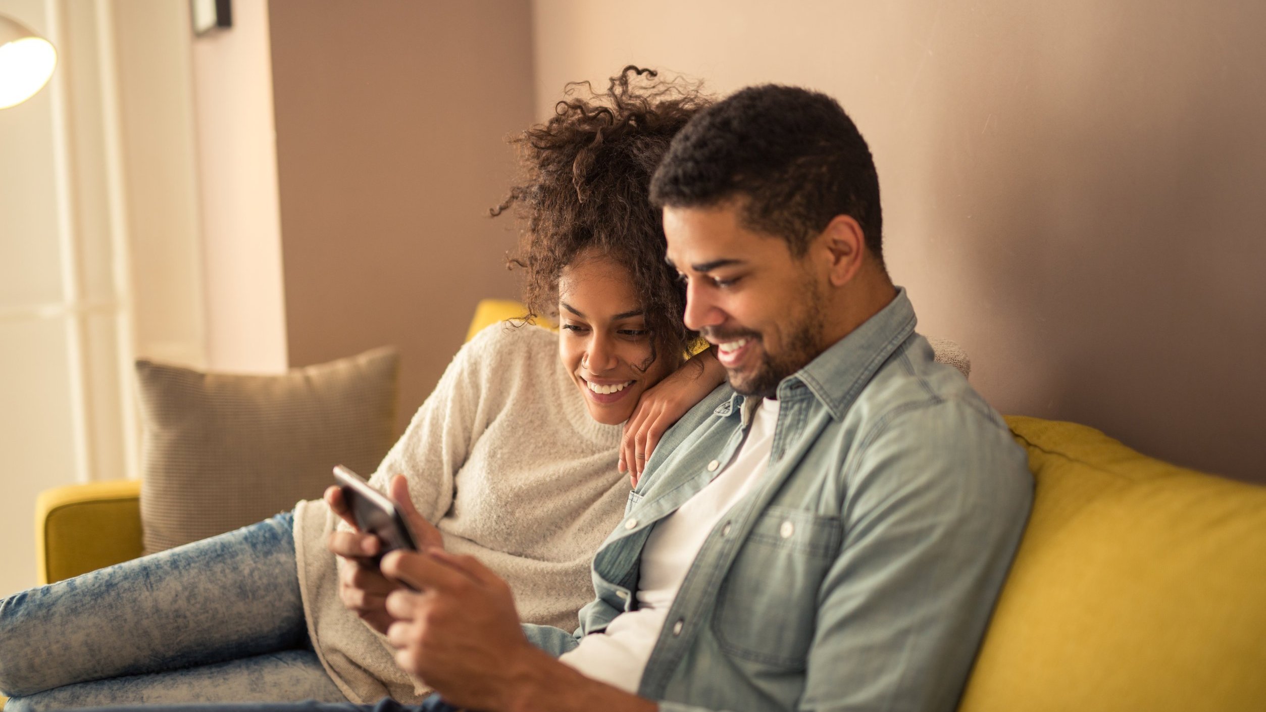 Casal sentado em sofá observa celular. Ambos sorriem.