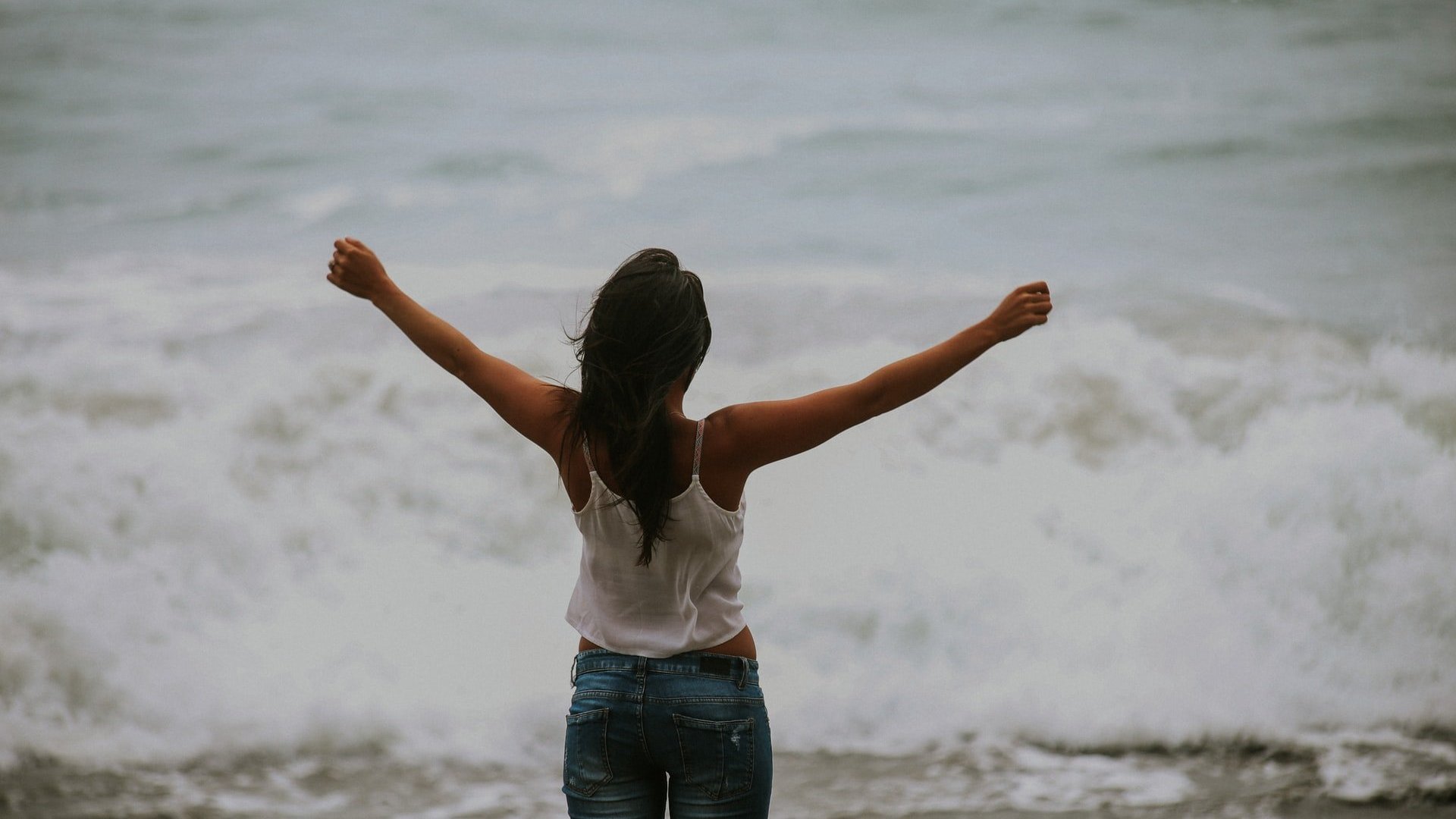 Ryan Moreno / Unsplash Mulher branca de costas na beira do mar com os braços abertos.