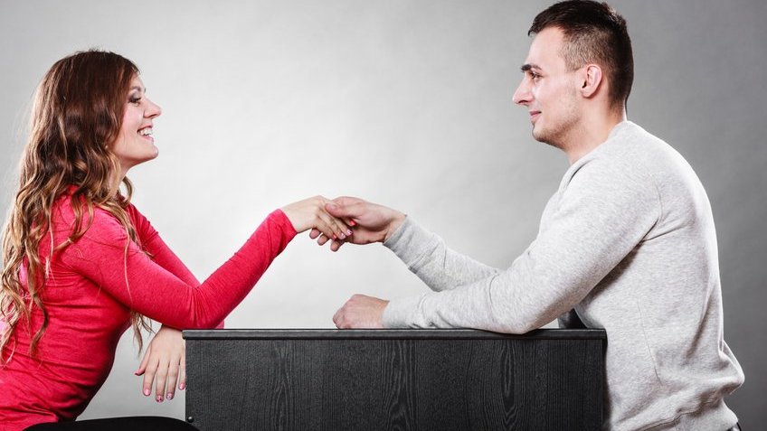 Homem e mulher dando aperto de mão, um de cada lado da mesa, em primeiro encontro