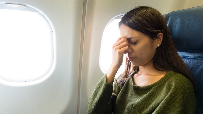 Mulher branca com a mão na testa sentada em uma poltrona de avião.