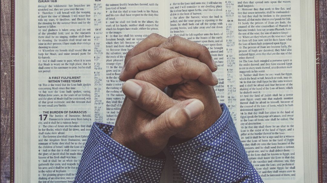 Duas mãos com dedos entrelaçados gesticulando um sinal de oração. As mãos estão debruçadas sobre uma bíblia aberta.