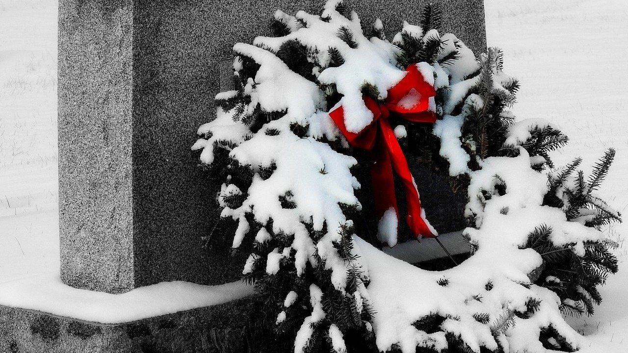 Uma guirlanda de natal coberta de neve e com um laço vermelho. Ela está repousada sobre uma lápide.
