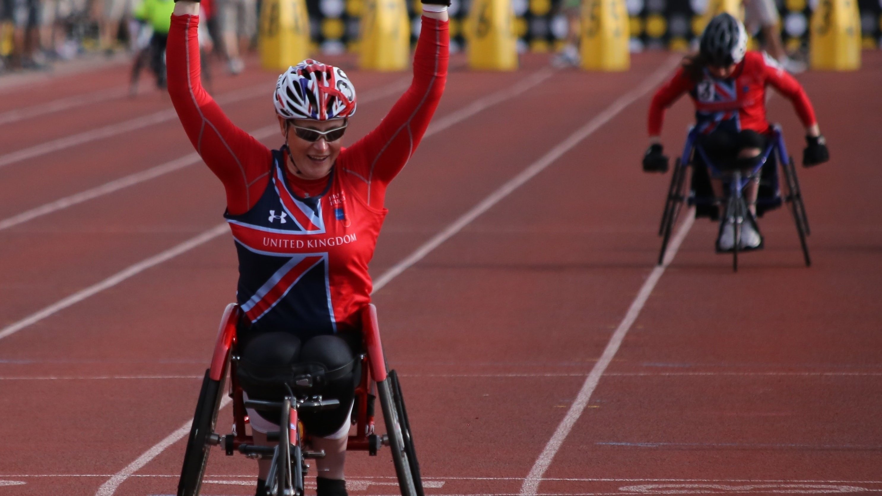Duas atletas paraolímpicas em uma corrida de cadeira de rodas. A primeira ergue os braços comemorando.