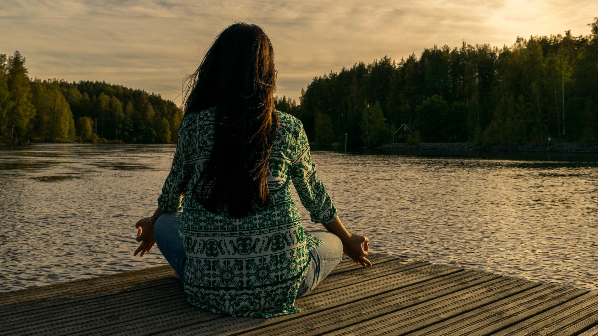Uma mulher de costas meditando em meio à natureza. Em frente, um rio e árvores.
