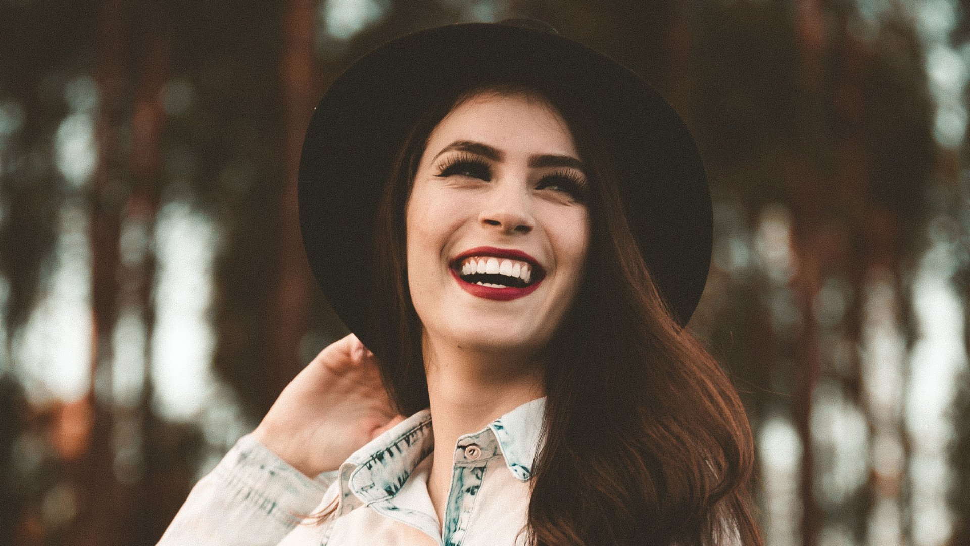 Uma mulher branca de chapéu preto e cabelos castanhos em uma floresta. Ela sorri intensamente.