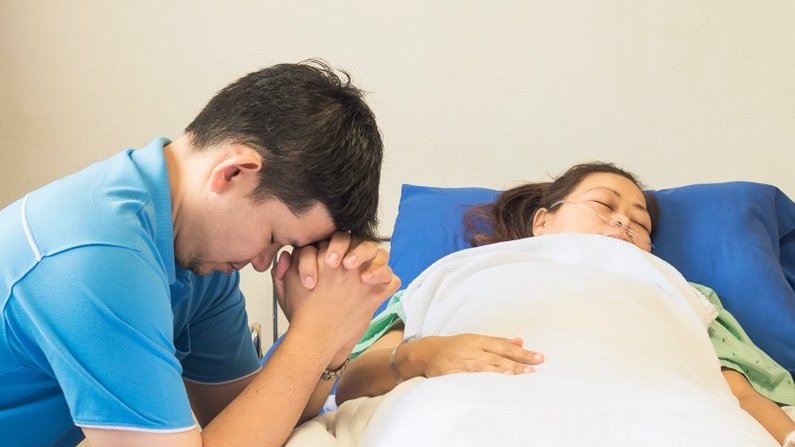 Homem faz uma oração na beira da cama onde mulher está deitada, doente