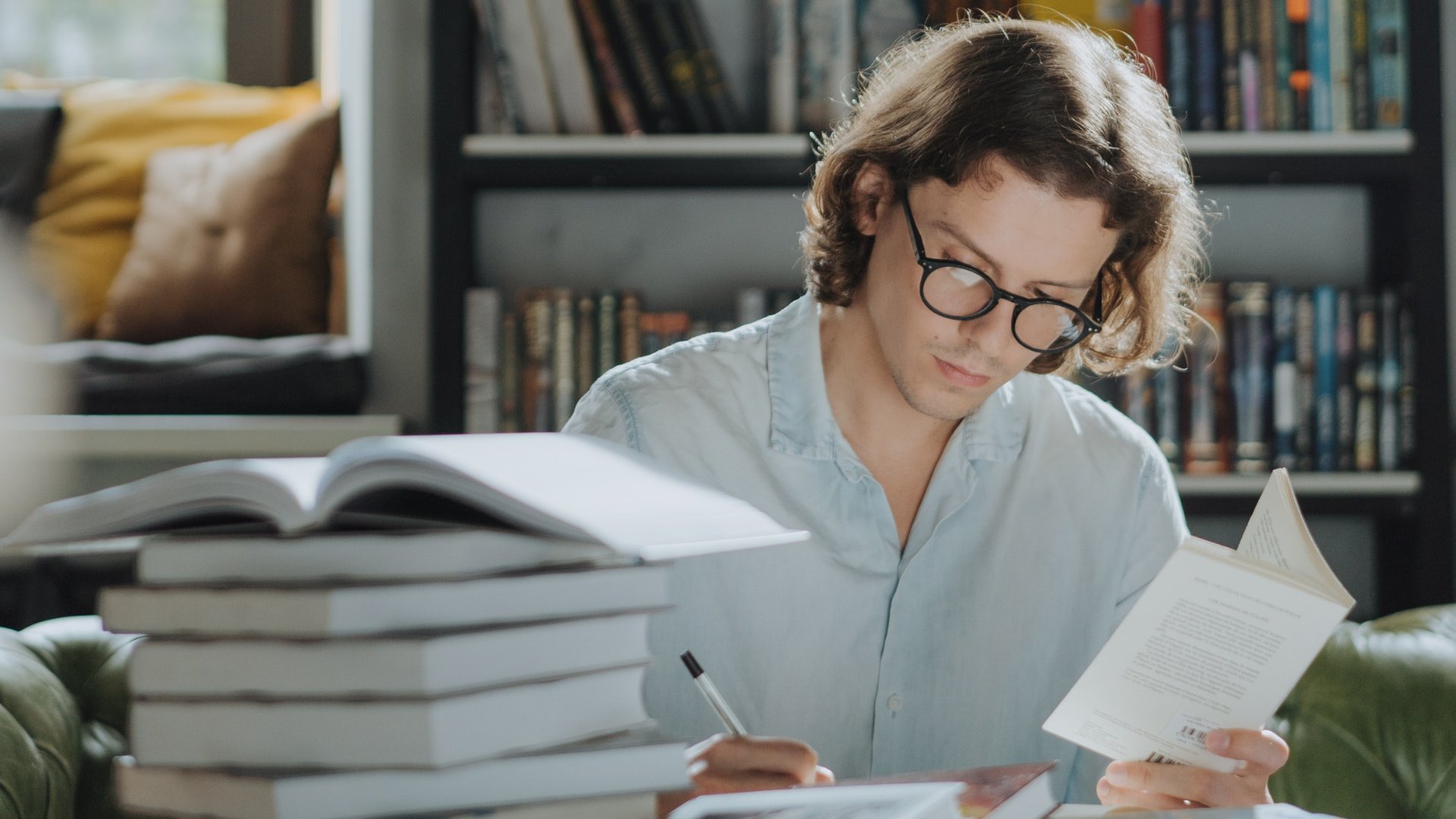 Um homem branco sentado. À sua frente, uma pilha de livros. Ele está escrevendo algo em um caderno.