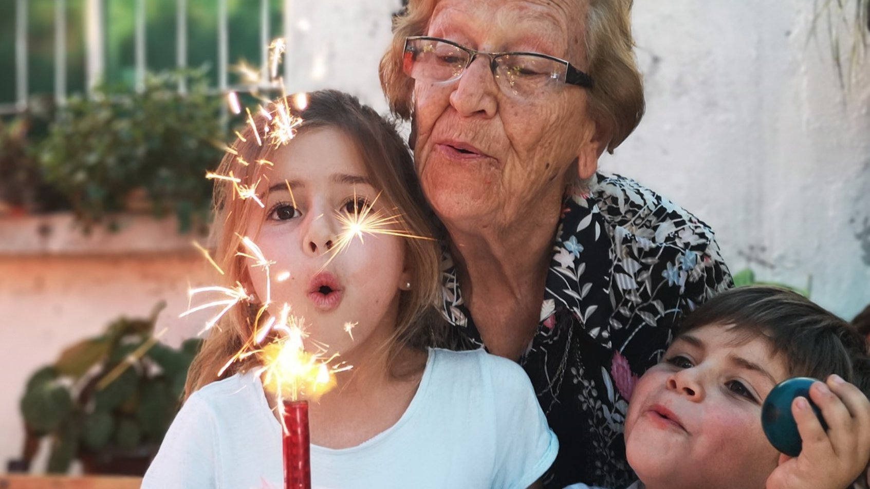 Uma mulher idosa, uma garota branca e um garoto branco comemorando um aniversário. À frente da garota, uma vela de aniversário.