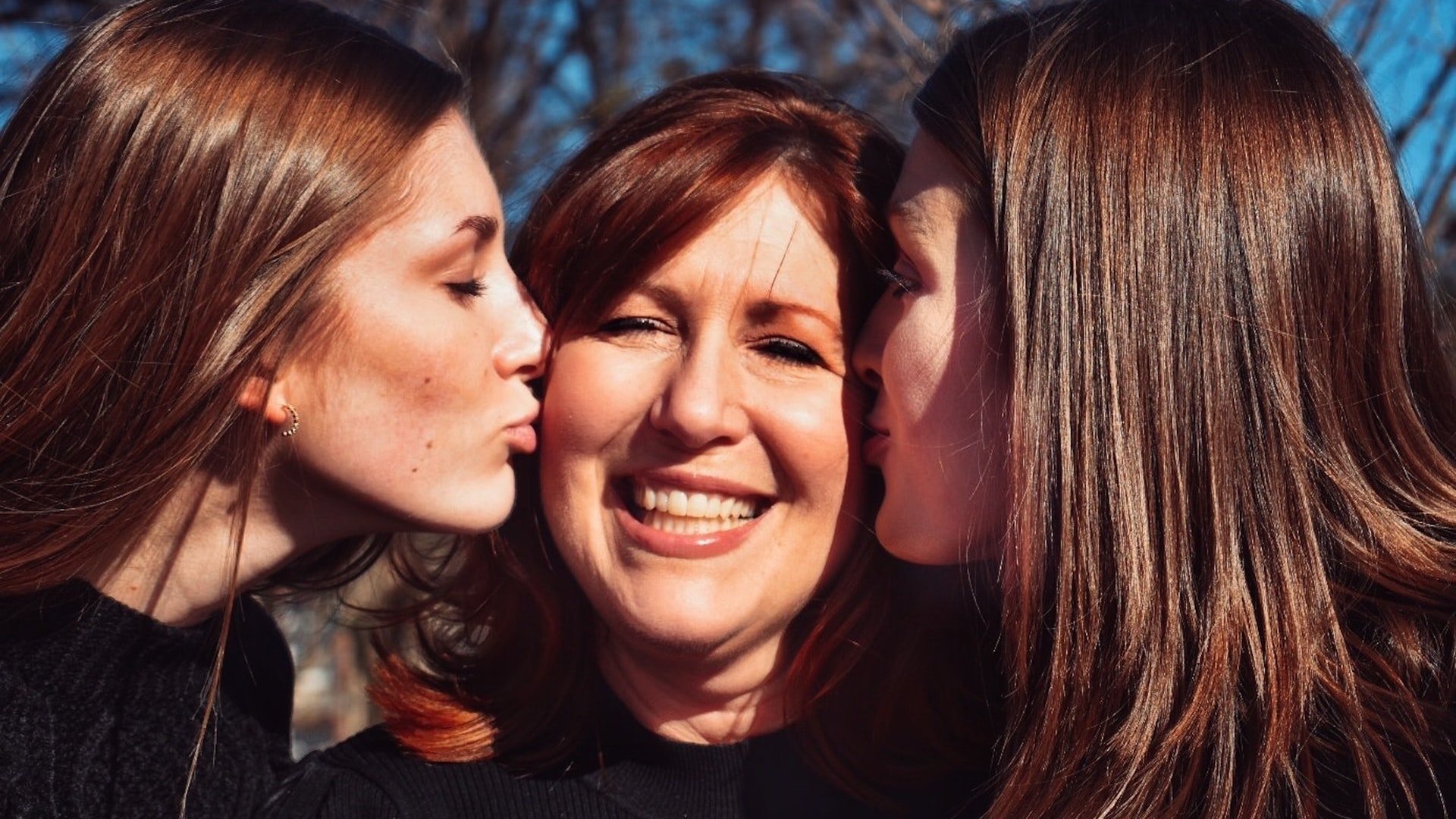 Uma mulher branca de cabelos castanhos sorrindo, ao centro. À esquerda, uma mulher dando-lhe um beijo no rosto, e à direita outra, fazendo a mesma coisa.