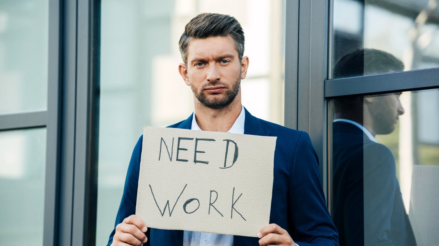 Um homem branco com um terno. Ele está segurando uma placa que diz 'need work'.