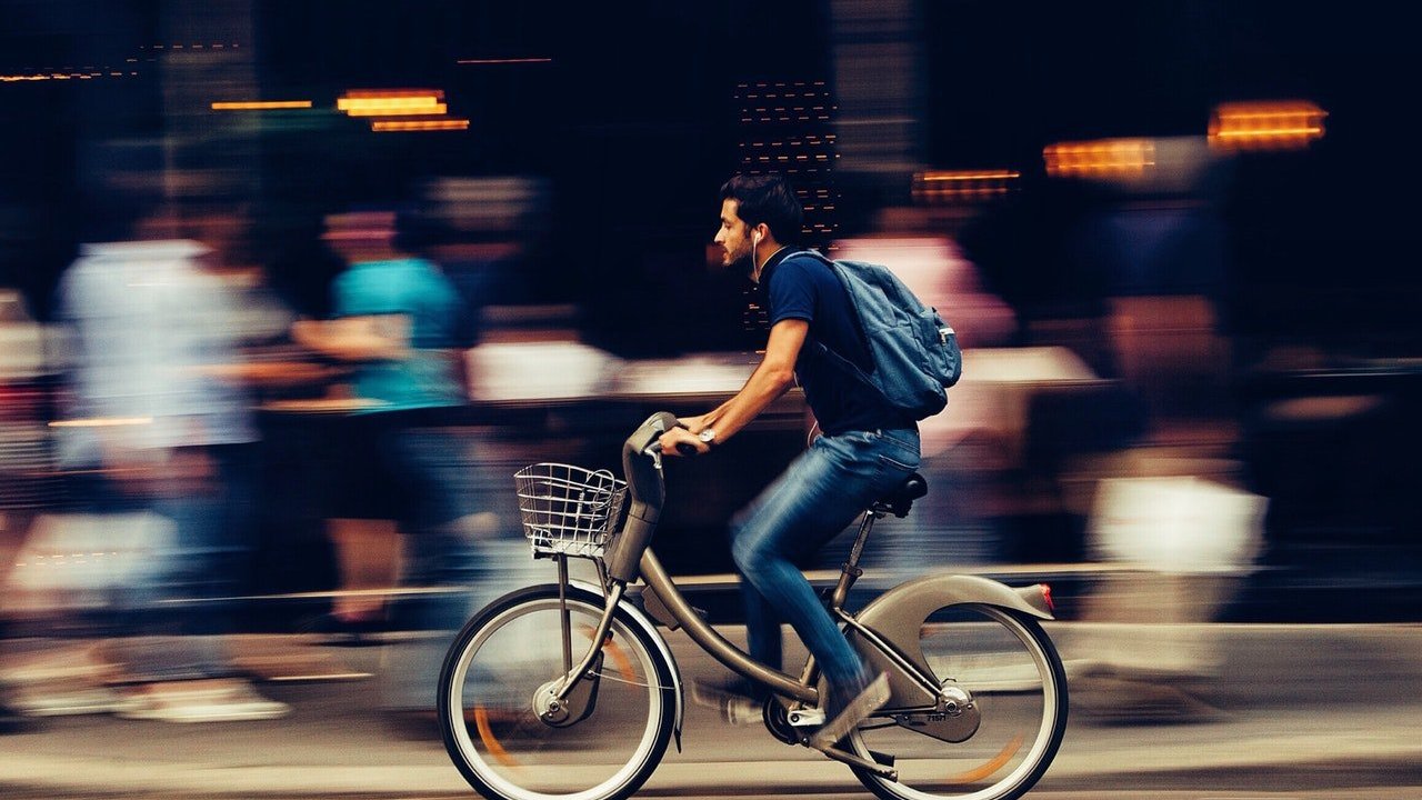 Um homem andando de bicicleta. Ao fundo, pessoas desfocadas.