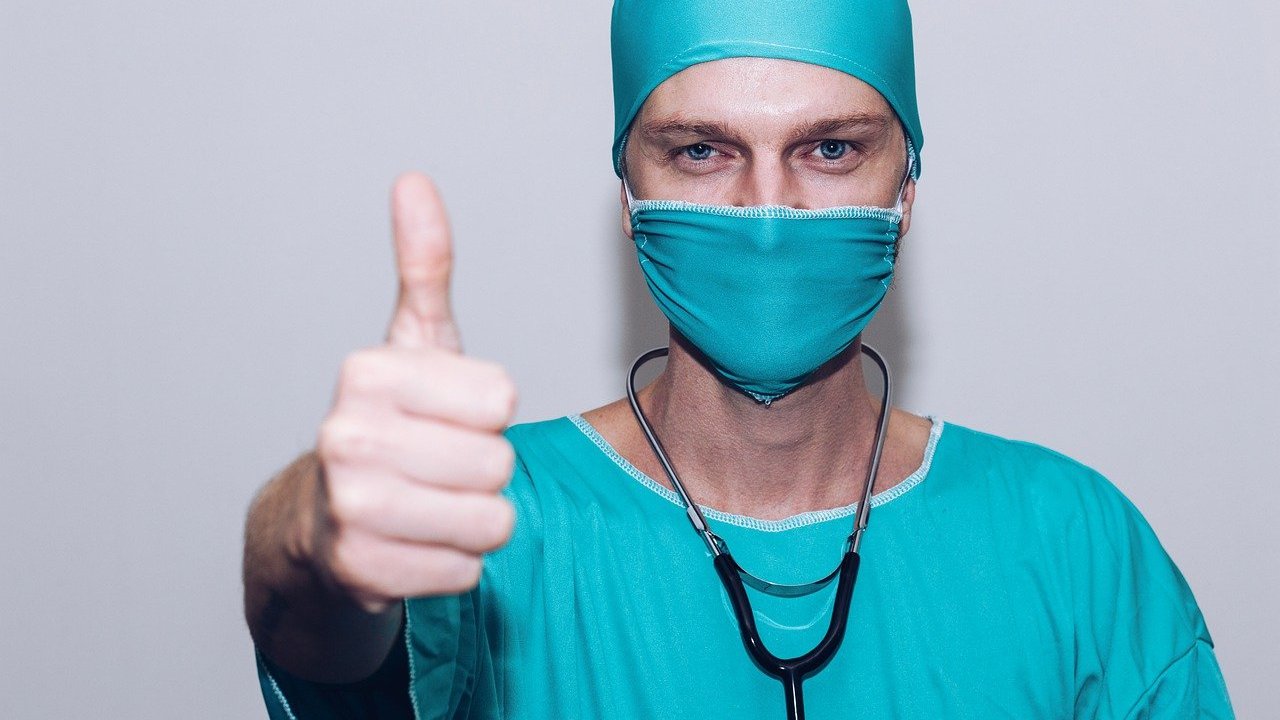 Um profissional da saúde trajado com uma máscara e roupa de cirurgião fazendo o gesto típico de joinha.