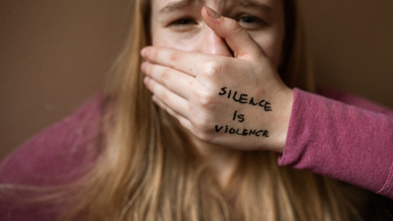 Uma mulher chorando e colocando a mão sobre a boca. Em sua mão está escrito silence is violence.