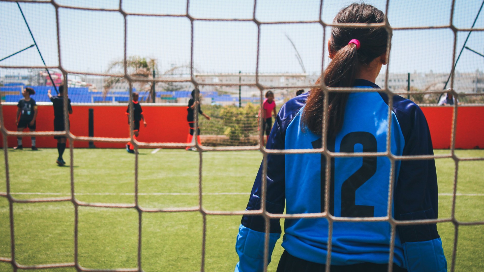 Uma mulher goleira, vista de costas. À frente dela, jogadores dispersos por um campo de futebol.
