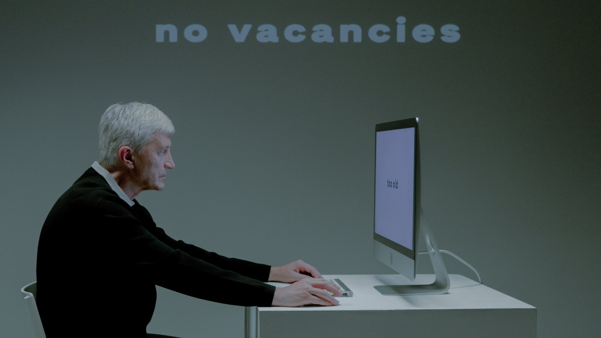 Um homem branco idoso em frente a um computador. No fundo, numa parede, a frase no 'vacancies', que em português significa 'não há vagas'.