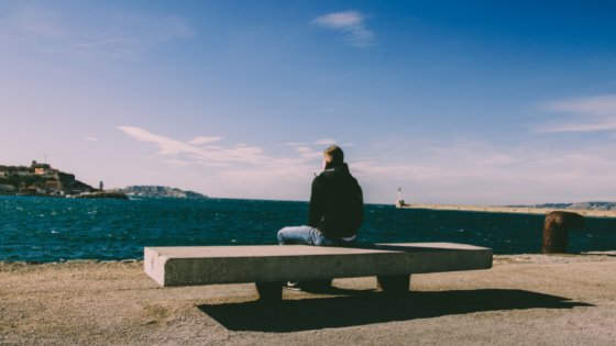 Homem sentado em banco de pedra de costas olhando para o mar