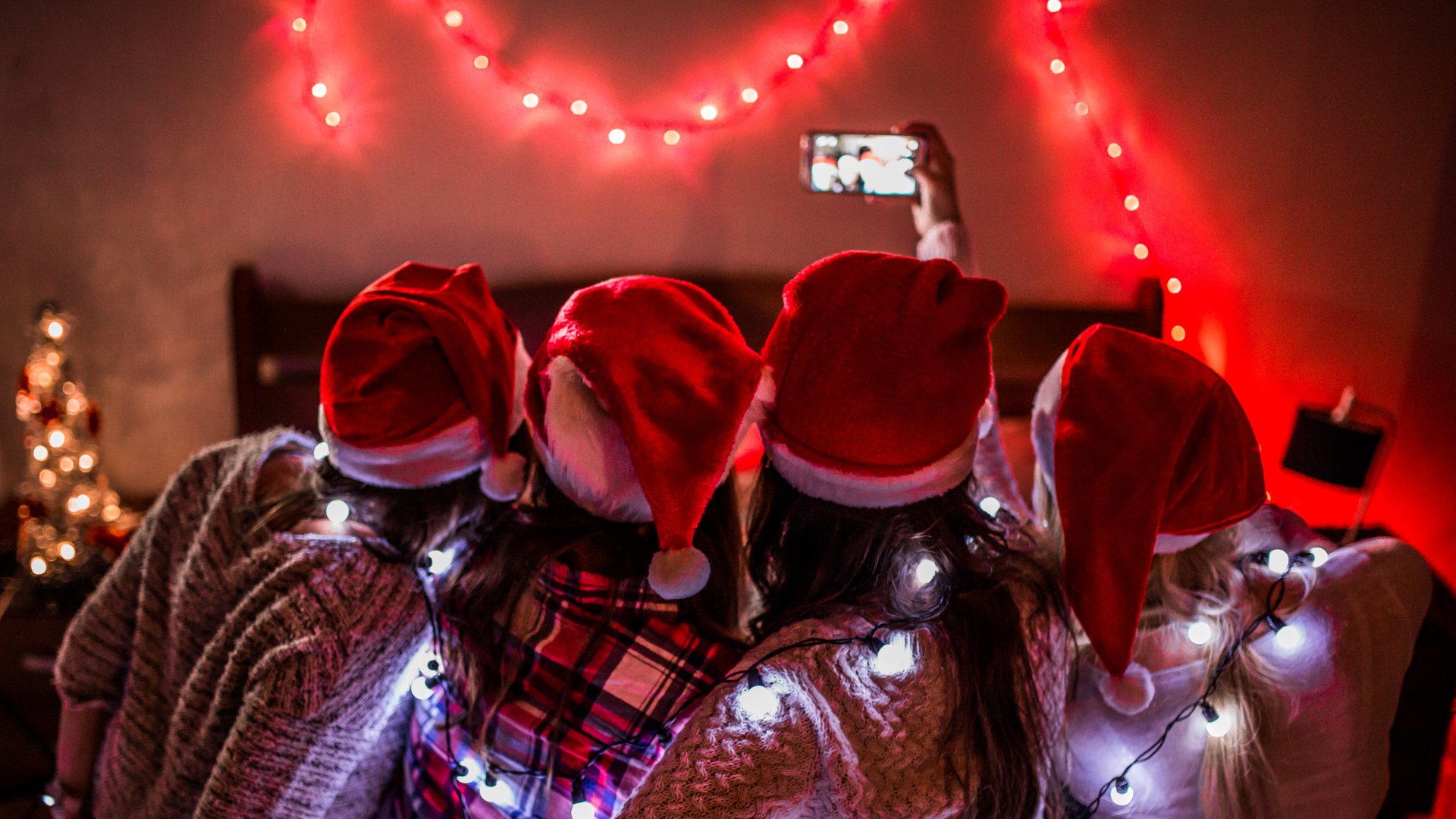 Pessoas juntas - abraçadas - vestindo gorros natalinos. Uma das pessoas ergue o celular, capturando uma selfie.