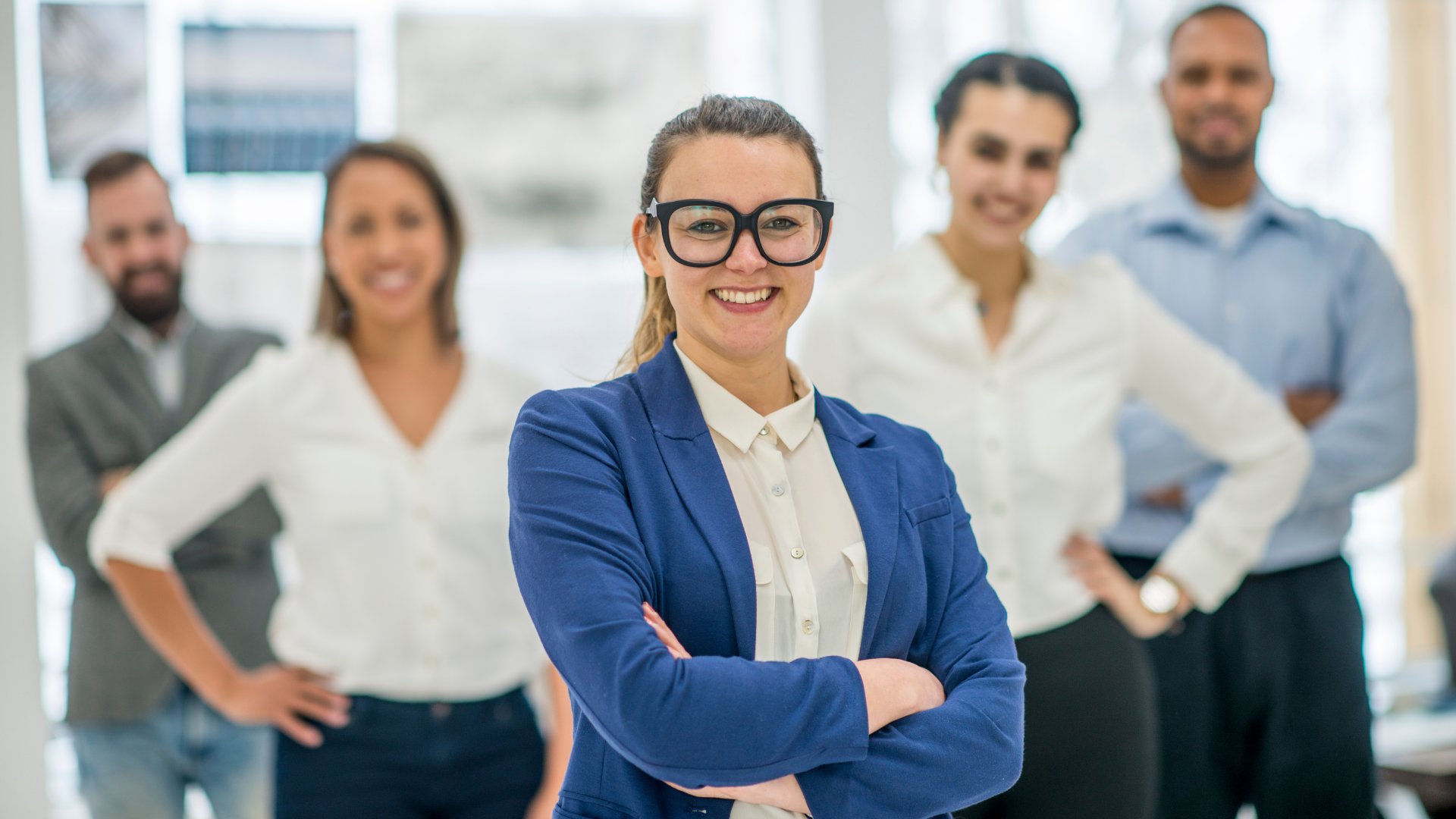 Em primeiro plano, uma mulher loira com um terno azul sorrindo. Em plano secundário e desfocado, homens e mulheres.