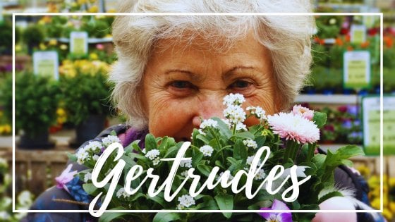 Mulher idosa branca sorrindo atrás de um arranjo de flores pequenas com escrito