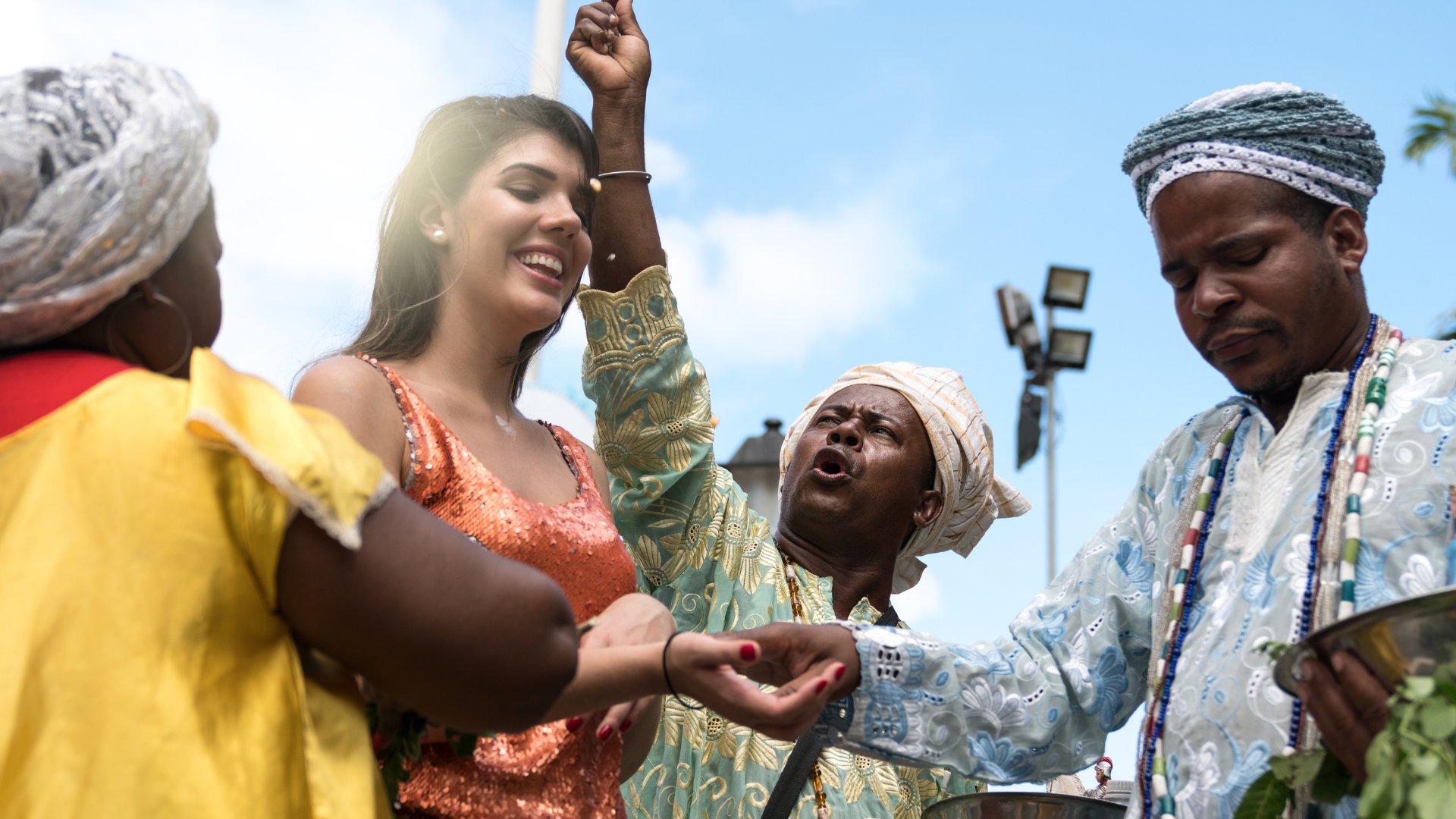 Uma mulher sorrindo. Em torno dela, homens, à direita e à esquerda, uma mulher benzendo-a.