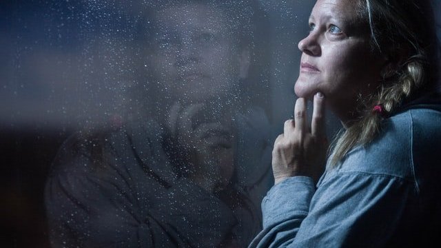 mulher com as mãos no queixo com cara de pensativa em frente a uma janela chuvosa