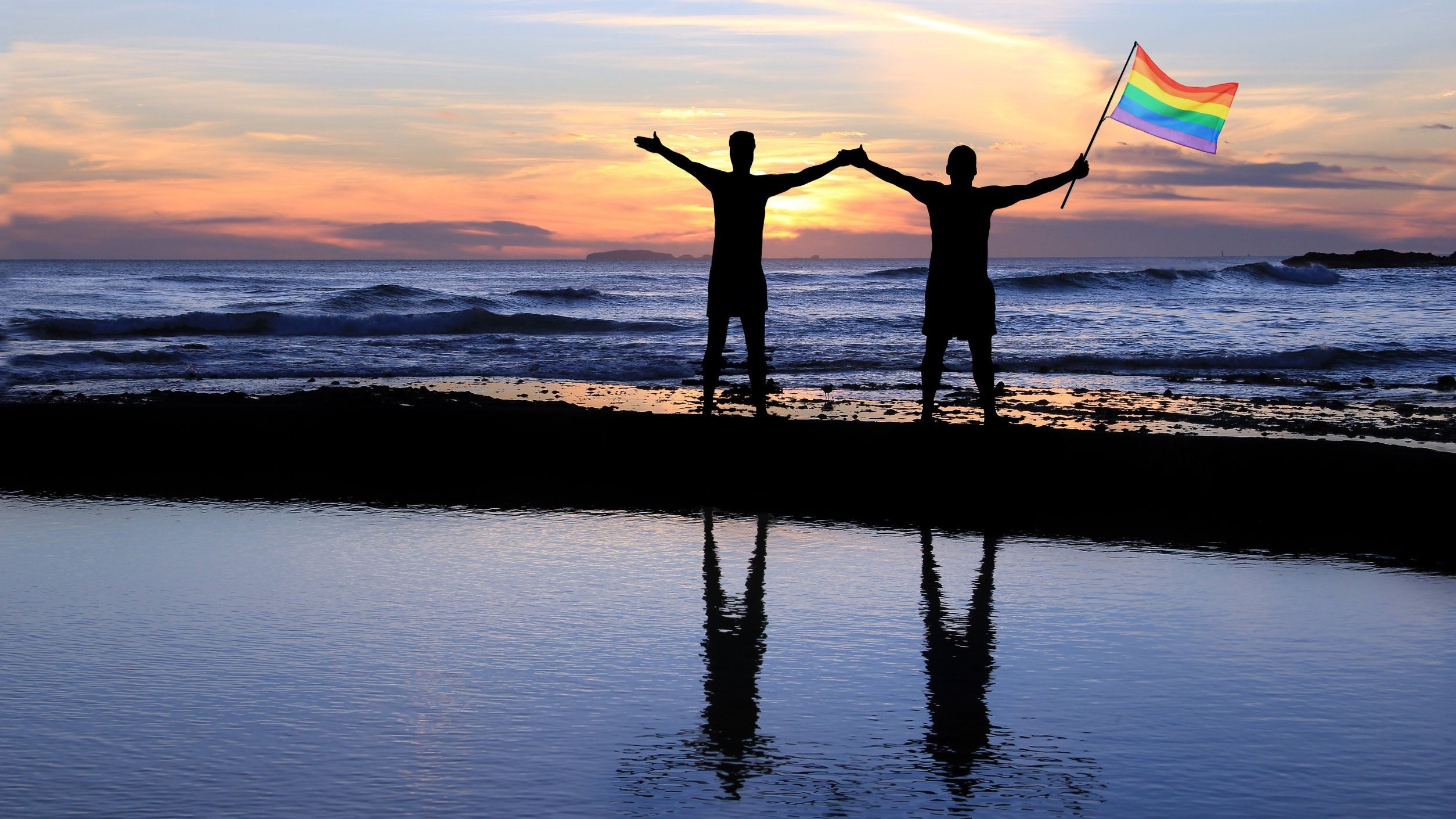 Casal de mão dadas na praia com a bandeira LGBTQI+