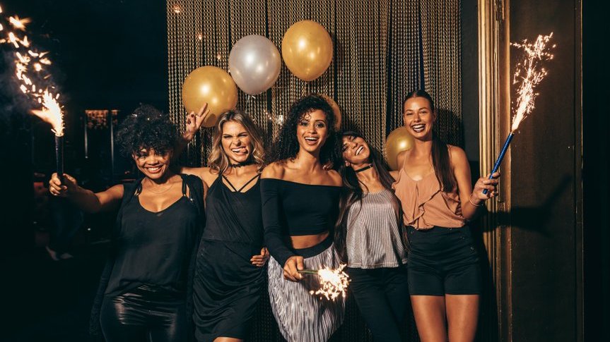 Cinco mulheres sorrindo abraçadas comemorando o ano novo em um ambiente aberto durante a noite