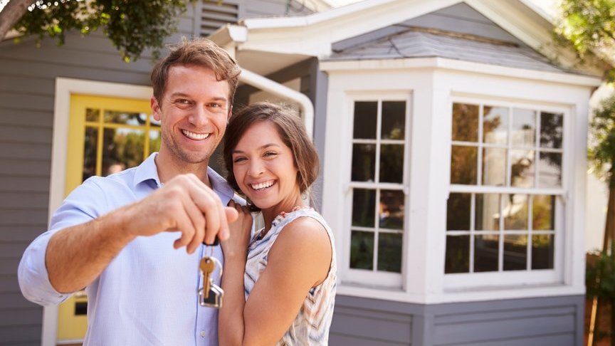 Homem e mulher abraçados e sorrindo em frente a uma casa enquanto ele segura as chaves do lar