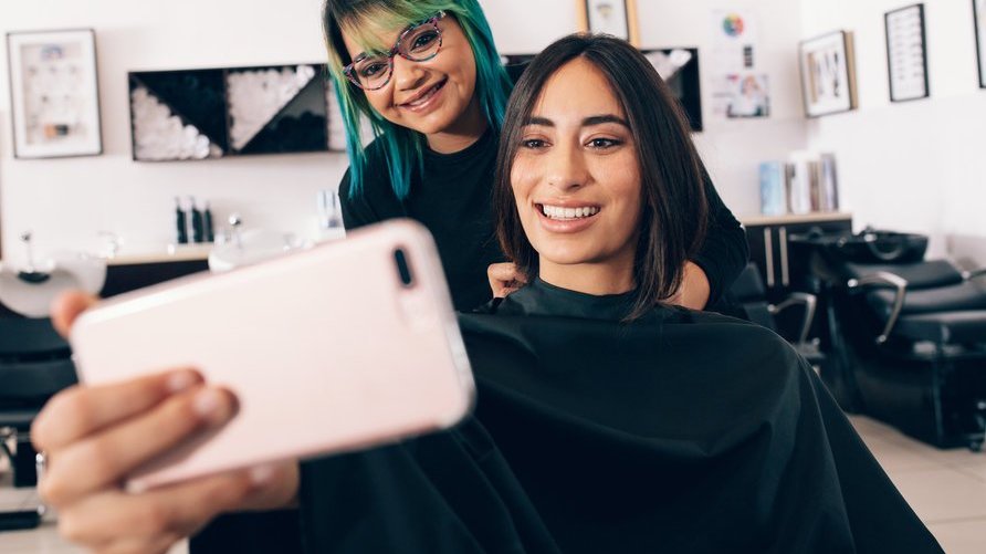 Mulher sentada tirando foto com outra mulher que está em pé cuidando do cabelo da primeira