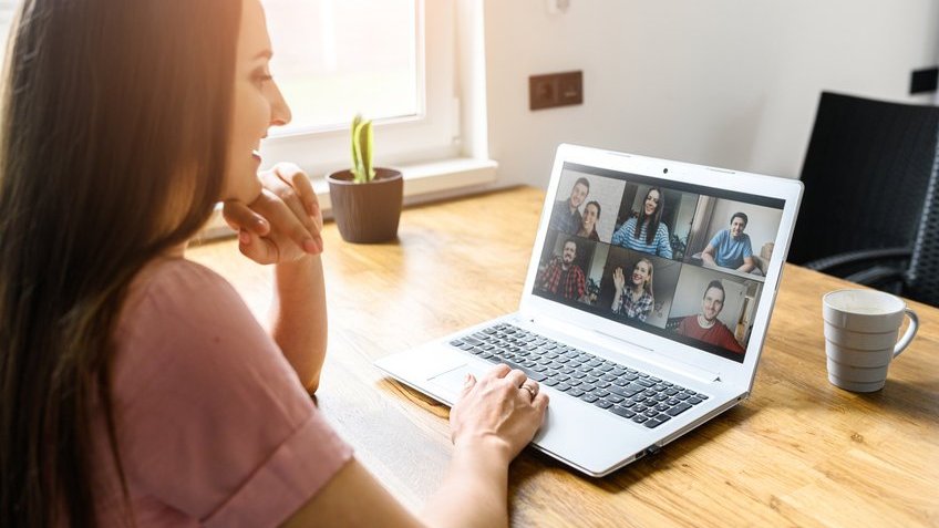 Mulher sentada sorrindo olhando a chamada de vídeo que aparece na tela do notebook que está em cima da mesa