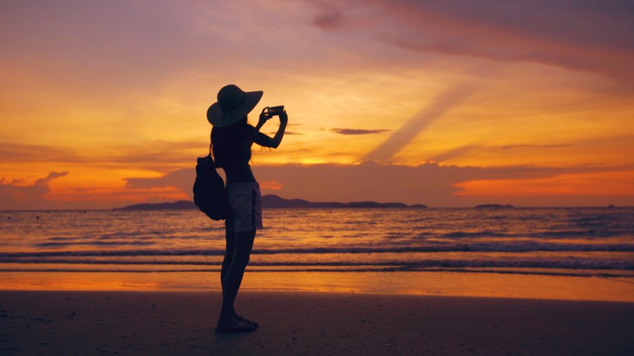 Mulher em pé na areia da praia tirando uma foto do crepúsculo