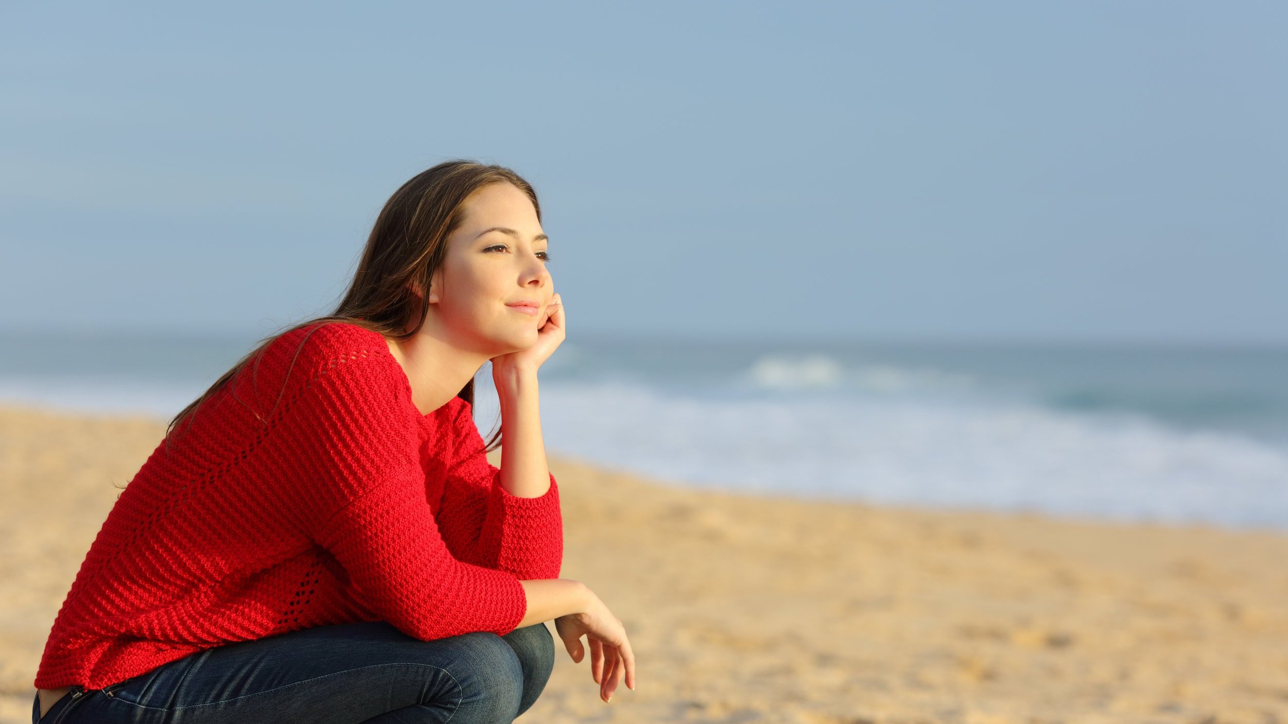 Mulher sentada na areia da praia olhando em direção ao horizonte com os braços apoiados no joelho