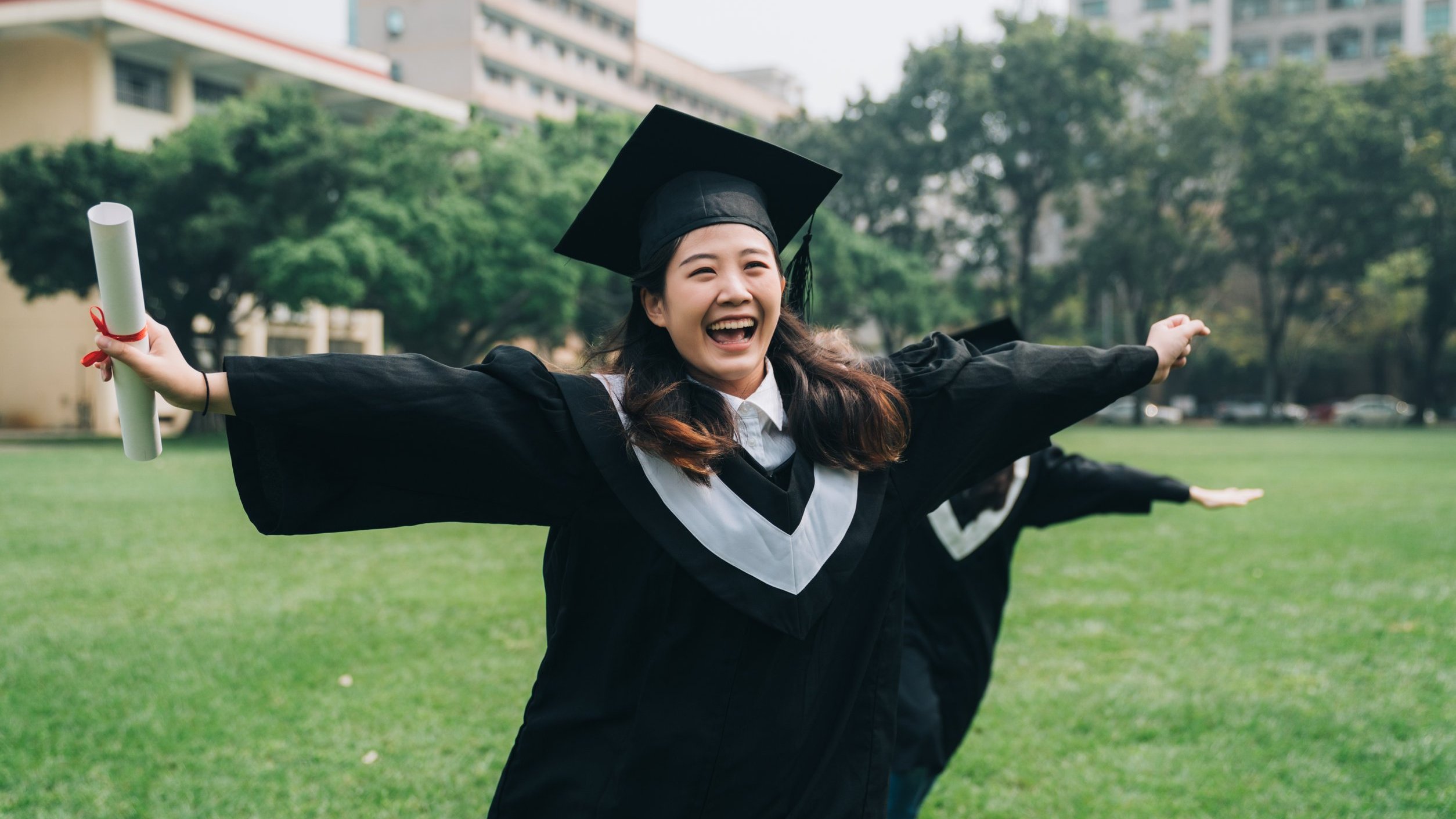 Mulher vestida de beca e capelo com os braços abertos comemorando segurando seu diploma